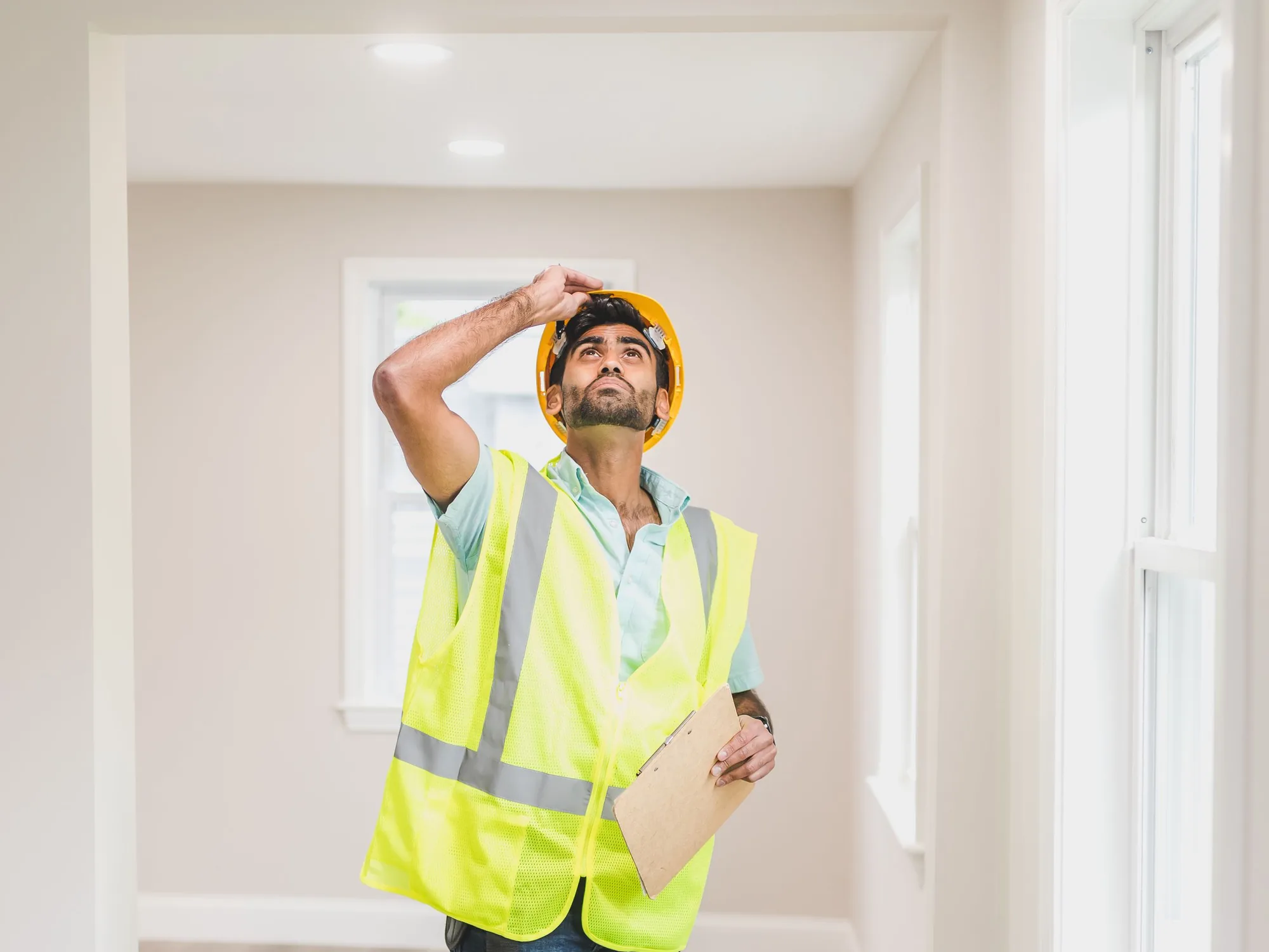 An inspector in a hard hat and safety vest looking up at a ceiling while holding a clipboard