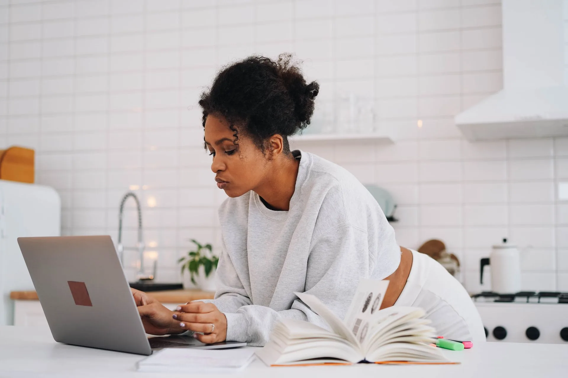 A woman researching on a laptop at her kitchen counter with an open book beside her