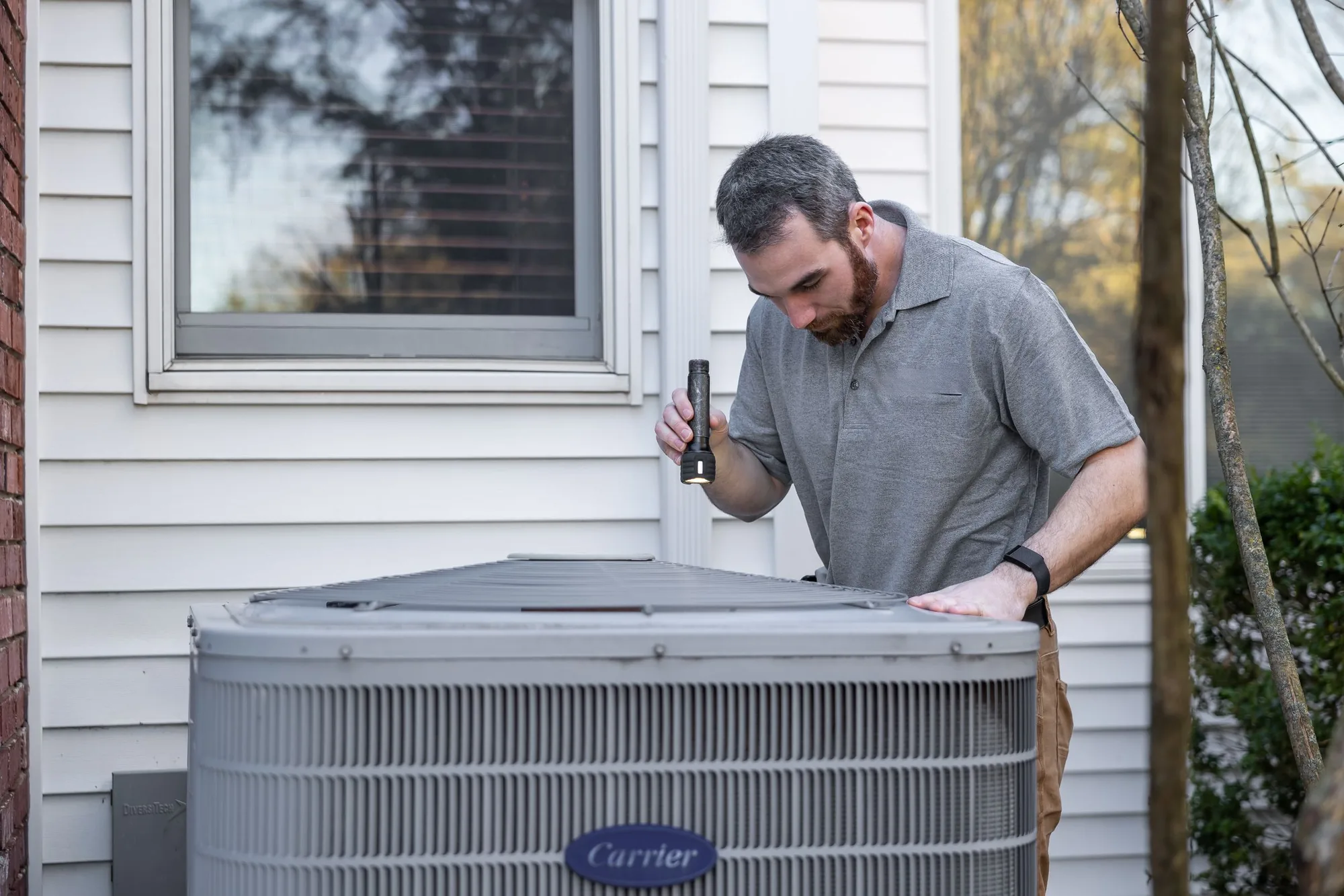 A technician with a flashlight inspecting an outdoor HVAC unit beside a home