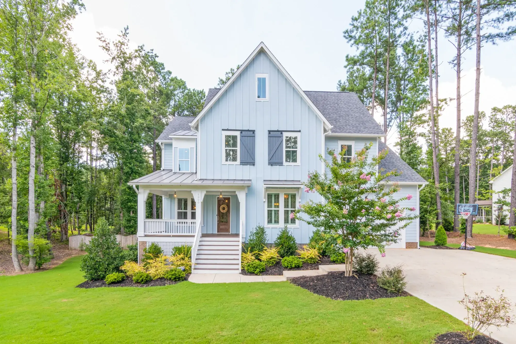 A light blue modern farmhouse-style home with a covered front porch, surrounded by tall pines