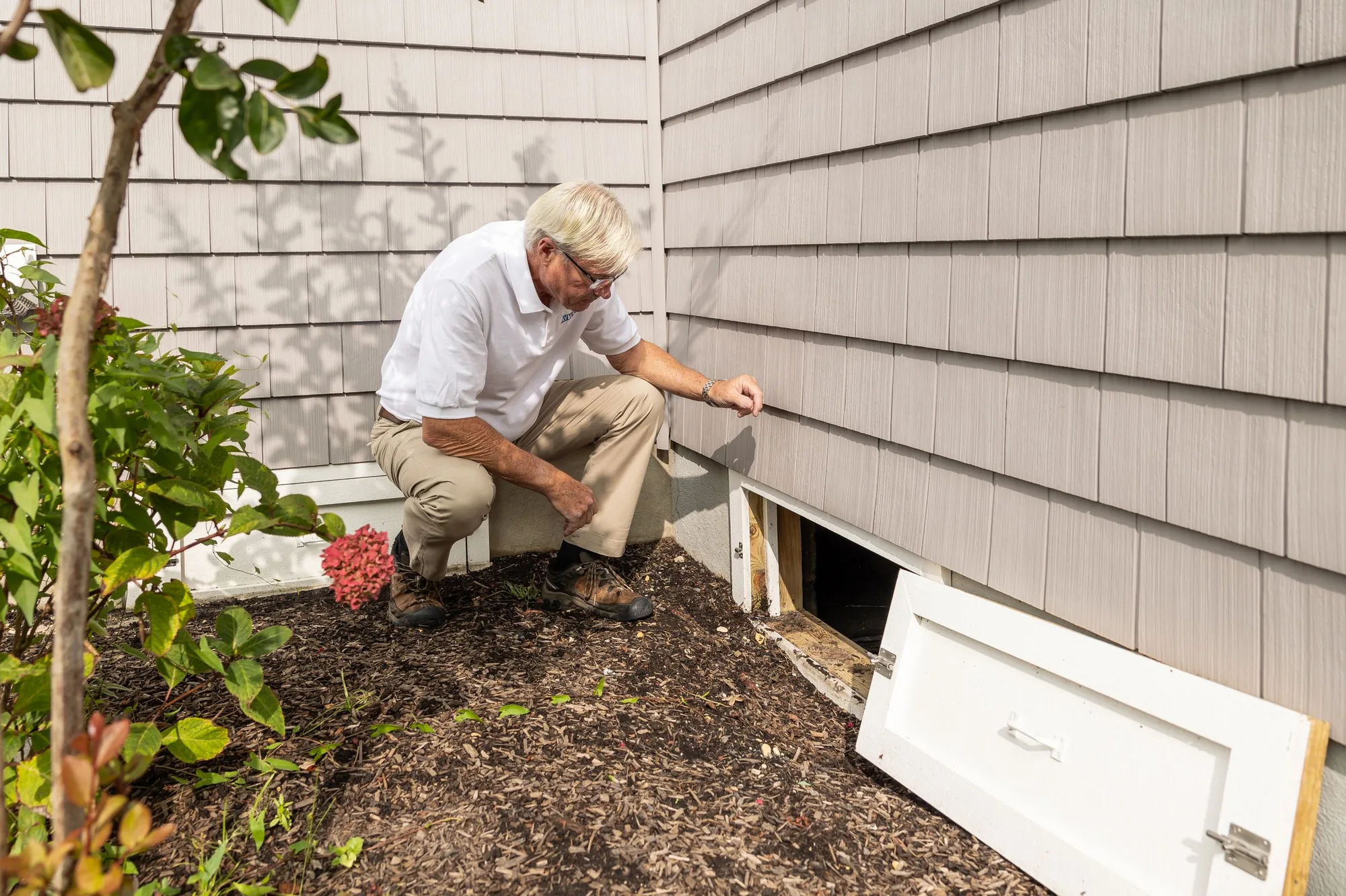 An inspector kneeling to examine an open crawlspace access door at the base of a home