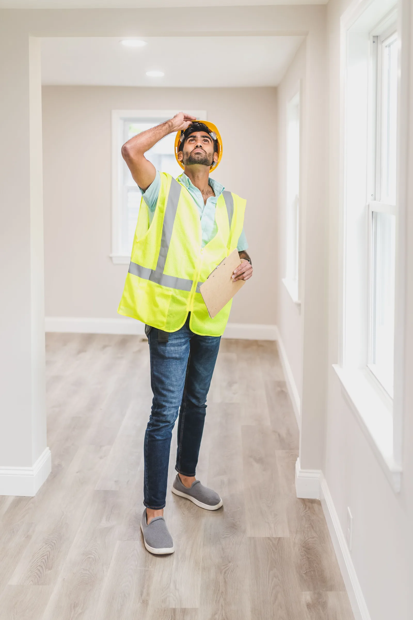 Independent inspector in a hard hat and safety vest examining a home's ceiling and walls with a clipboard during a mold assessment